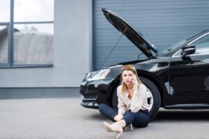 Woman sitting on the ground next to a broken-down black car with an open hood, making a phone call for roadside assistance.